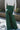 Back view of model wearing the Nothing But Love Pants, that have dark green fabric with inverted front pleating, an attached tie belt, wide flowy legs, and a side zipper. Worn with white top.