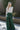 Full body back view of model wearing the Nothing But Love Pants, that have dark green fabric with inverted front pleating, an attached tie belt, wide flowy legs, and a side zipper. Worn with white top.