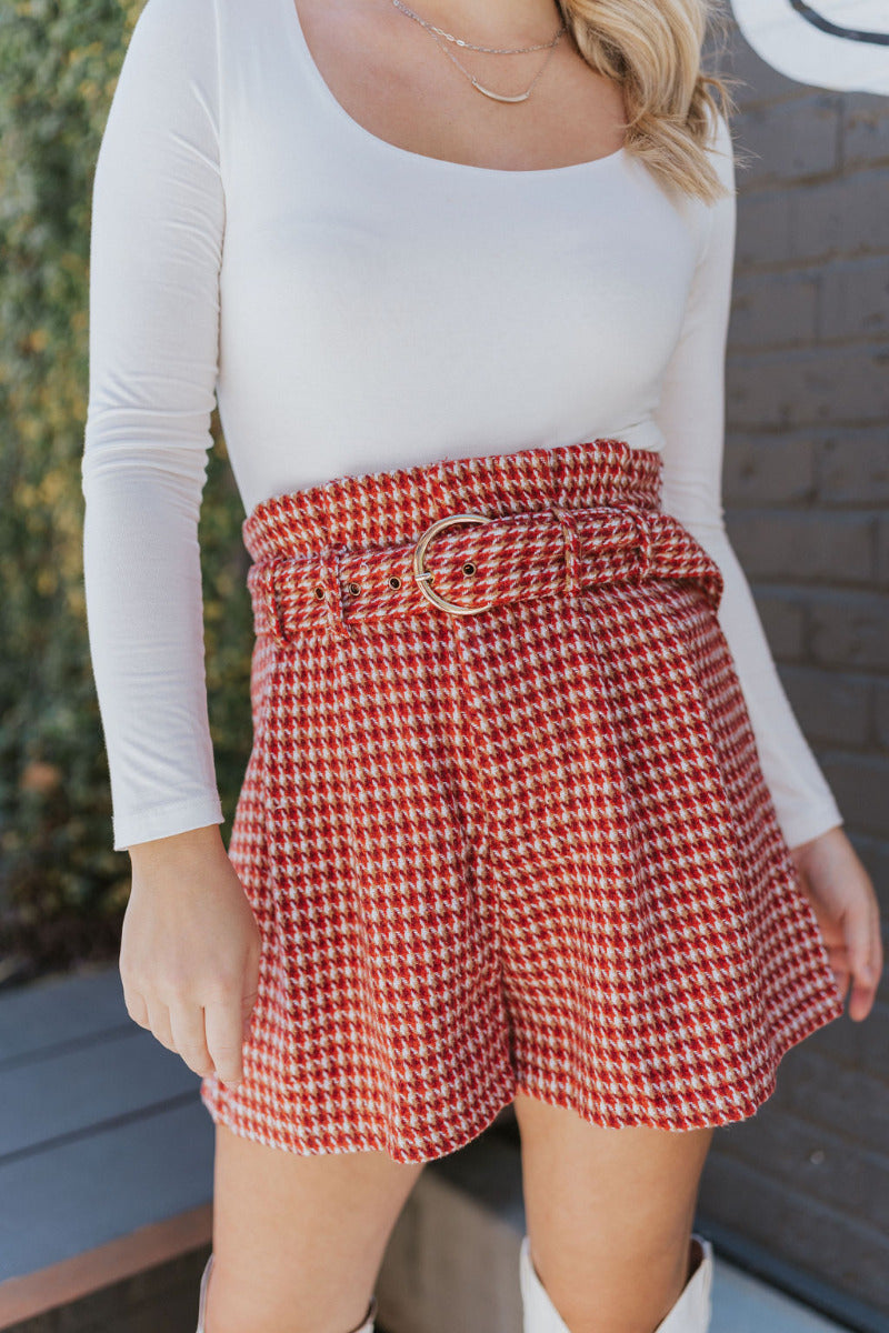 Close front view of model wearing the Around The World Tweed Shorts, that have a rust-colored tweed fabric, a matching belt with a gold buckle, a front zipper and hook closure, and a high-rise waist. Worn with white top.