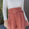 Close front view of model wearing the Around The World Tweed Shorts, that have a rust-colored tweed fabric, a matching belt with a gold buckle, a front zipper and hook closure, and a high-rise waist. Worn with white top.