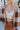 Close front view of model wearing the Check Mate Cardigan, that has a brown, tan, and ivory checker pattern, a button-up front with four buttons, balloon sleeves, and ribbed trim. Worn unbuttoned over white brami.
