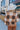 Side view of model wearing the Check Mate Cardigan, that has a brown, tan, and ivory checker pattern, a button-up front with four buttons, balloon sleeves, and ribbed trim. Worn unbuttoned over white brami.