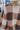 Close back view of model wearing the Check Mate Cardigan, that has a brown, tan, and ivory checker pattern, a button-up front with four buttons, balloon sleeves, and ribbed trim. Worn unbuttoned over white brami.