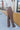 Side view of model wearing the Don't Wait Up Pants, that have brown woven fabric with front pleating, a front zipper and hook closure, side pockets, and faux back pockets