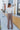 Full body back view of model wearing the Don't Wait Up Pants, that have brown woven fabric with front pleating, a front zipper and hook closure, side pockets, and faux back pockets