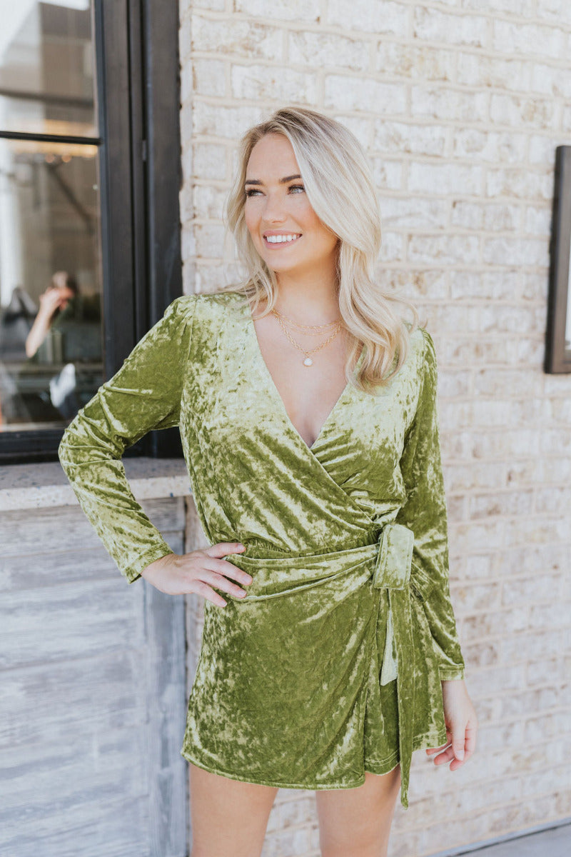 Front view of model wearing the Supernova Velvet Romper in Green, which has light green crushed velvet, a surplice neckline, a front skirt overlay with a side tie, and long fitted sleeves