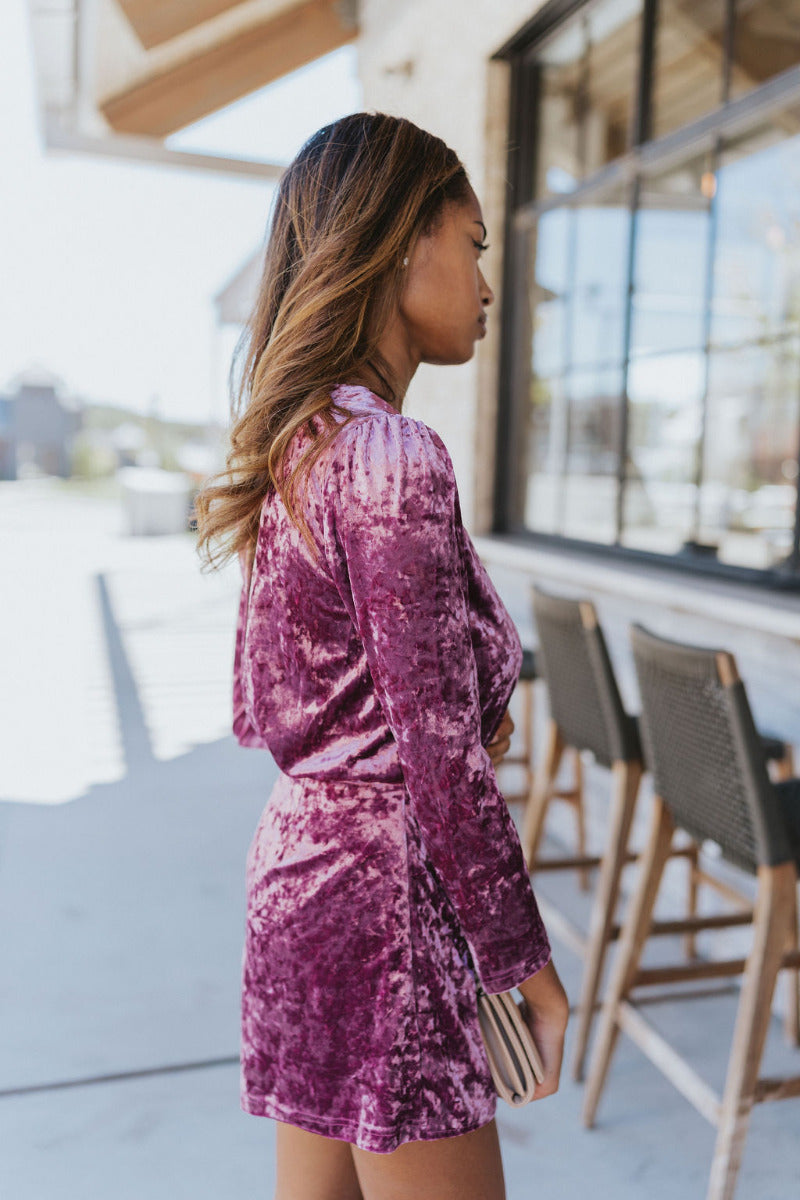 Side view of model wearing the Supernova Velvet Romper in Plum, which features light purple crushed velvet, a surplice neckline, a front skirt overlay with a side tie, and long fitted sleeves