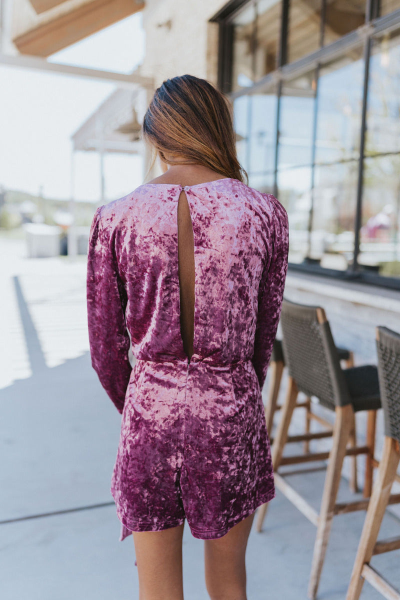 Back view of model wearing the Supernova Velvet Romper in Plum, which features light purple crushed velvet, a surplice neckline, a front skirt overlay with a side tie, and long fitted sleeves