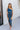 Front view of model wearing the Athena Midi Dress, that has blue satin fabric, a one-shoulder neckline with a braided back strap, a back cutout, and an asymmetrical midi hem.
