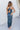Back view of model wearing the Athena Midi Dress, that has blue satin fabric, a one-shoulder neckline with a braided back strap, a back cutout, and an asymmetrical midi hem.