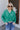 Front view of model wearing the Must Be True Top, which features green fabric, a button-up front with a tucked twist detail, long sleeves, and a collared neckline.