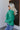 Side view of model wearing the Must Be True Top, which features green fabric, a button-up front with a tucked twist detail, long sleeves, and a collared neckline.