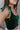 Close-up side view of model wearing the Luna Velvet Bodysuit in Green, which features velvet fabric, satin tie straps, and a cheeky bottom. Worn with jeans.