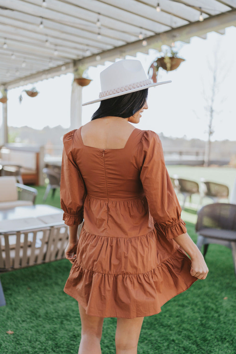 Back view of The Caramel Apple Dress features camel coloring fabric, tiered detailing, square neckline, mini length, back zipper and 1/2 puff sleeve with elastic hem