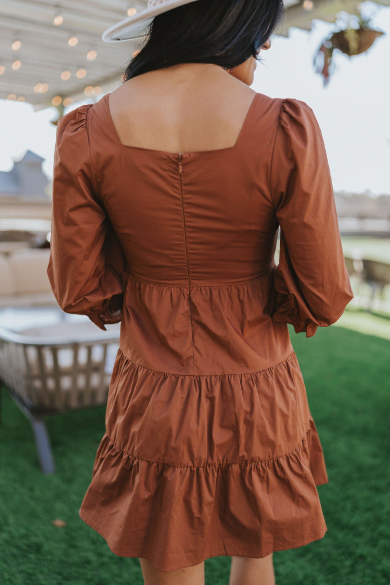 Close up back view of The Caramel Apple Dress features camel coloring fabric, tiered detailing, square neckline, mini length, back zipper and 1/2 puff sleeve with elastic hem