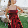 Front view of The Holiday Plans Dress features burgundy sheen fabric, babydoll style, square neckline with adjustable spaghetti straps, mini length and a bow on the back.