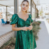 Front view of The All That Glistens Dress features metallic green fabric, bow tie on front chest, scooped neckline, mini length, short puff sleeves, and elastic waistline 