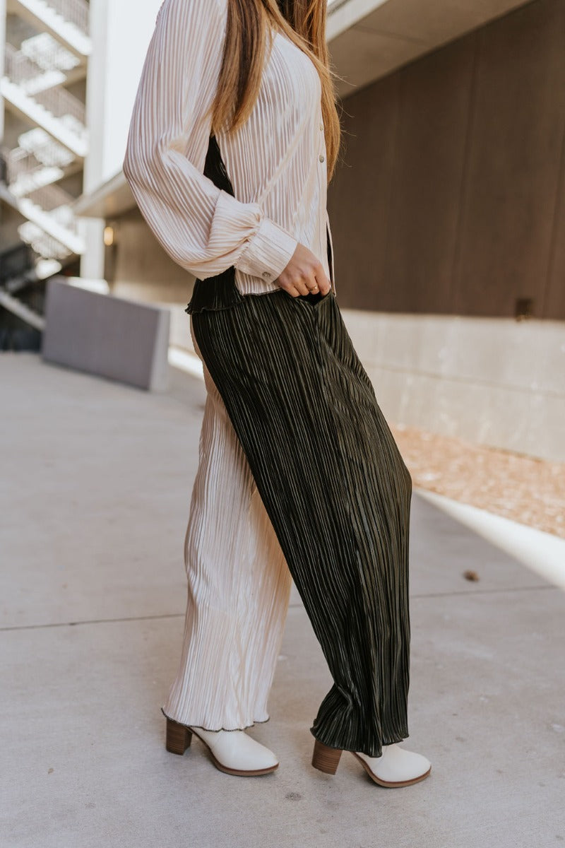 Side view of model wearing the Block It Out Pants that have olive and cream color block coloring, plisse fabric, two front pockets, and elastic waistband, and flare wide legs with lettuce hems.