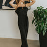 Front view of The Fresh Eyes Jumpsuit features black fabric, sweetheart neckline with a v wire, feather details, adjustable spaghetti straps, tie around the waist, two front pockets, back zipper, open back and flare pant leg.
