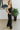 Side view of The Fresh Eyes Jumpsuit features black fabric, sweetheart neckline with a v wire, feather details, adjustable spaghetti straps, tie around the waist, two front pockets, back zipper, open back and flare pant leg.