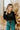 Upper front view of model wearing the Let's Chat Pants in Emerald that have emerald green fabric, a seam down each front leg, a slight v waistline, a back zipper closure, and wide legs