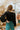 Back view of model wearing the The Late Nights Velvet Top that has  black velvet fabric, a cropped waist, ruched front detailing, a  v neckline, and long sleeves with elastic wrists.
