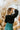 Back view of model wearing the The Late Nights Velvet Top that has  black velvet fabric, a cropped waist, ruched front detailing, a  v neckline, and long sleeves with elastic wrists.
