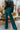 Back view of model wearing the Let's Chat Pants in Emerald that have emerald green fabric, a seam down each front leg, a slight v waistline, a back zipper closure, and wide legs