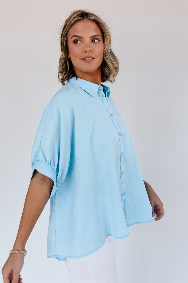 Side view of model wearing the Grace Washed Light Blue Short Sleeve Top which features light washed blue tencel fabric, monochrome front button up closure, collared neckline and short sleeves.