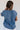 Back view of model wearing the Allison Washed Denim Ruffle Short Sleeve Top which features washed denim fabric, slight slits on each side, round collar with v-neckline and short sleeves with ruffle hem.