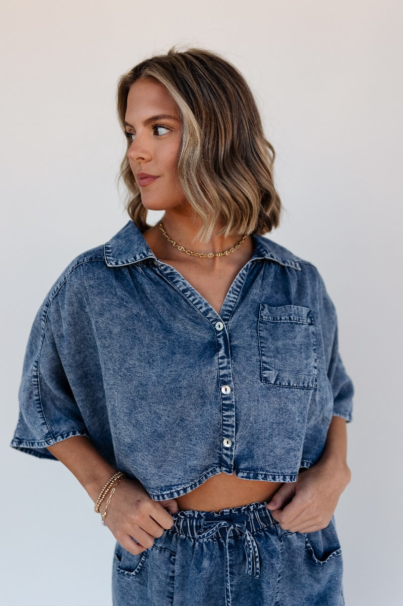 Front view of model wearing the Jade Washed Denim Short Sleeve Buttoned Top which features washed denim tencel fabric, cropped waist, pearlescent button up front closure, left front chest pocket, collared neckline and short sleeves.