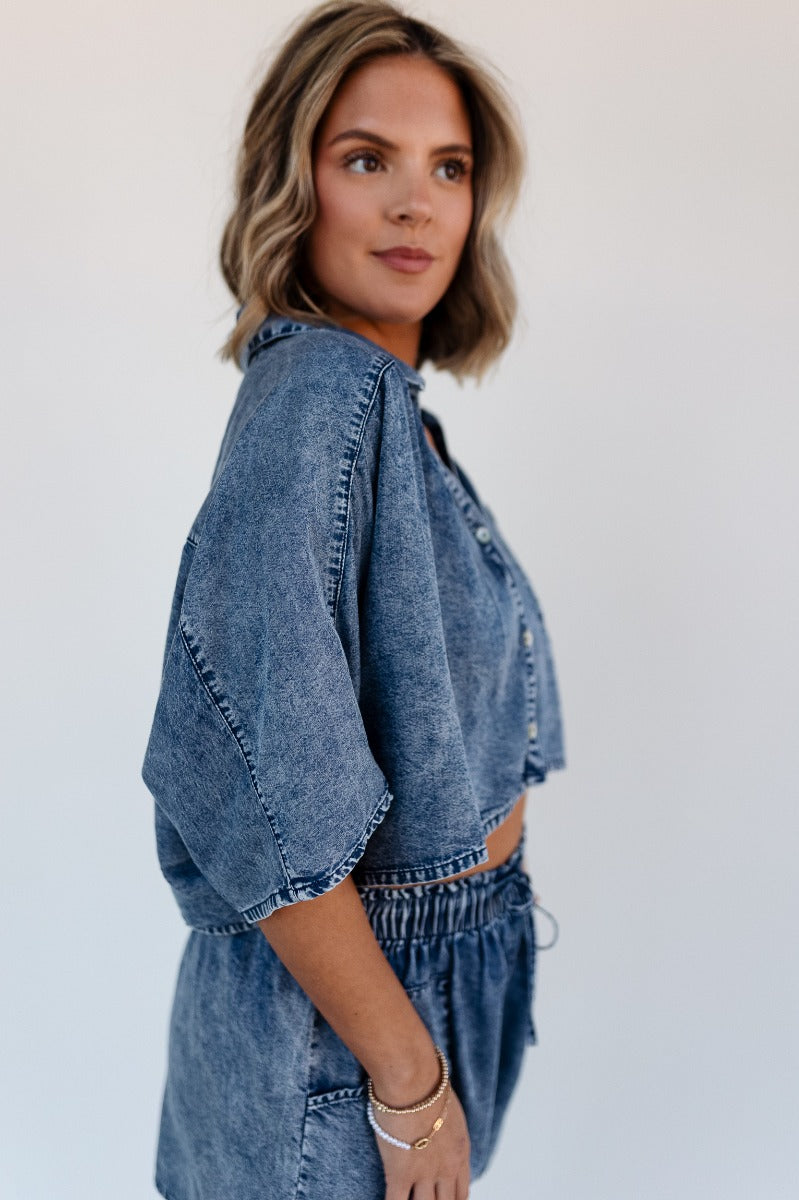 SIde view of model wearing the Jade Washed Denim Short Sleeve Buttoned Top which features washed denim tencel fabric, cropped waist, pearlescent button up front closure, left front chest pocket, collared neckline and short sleeves.