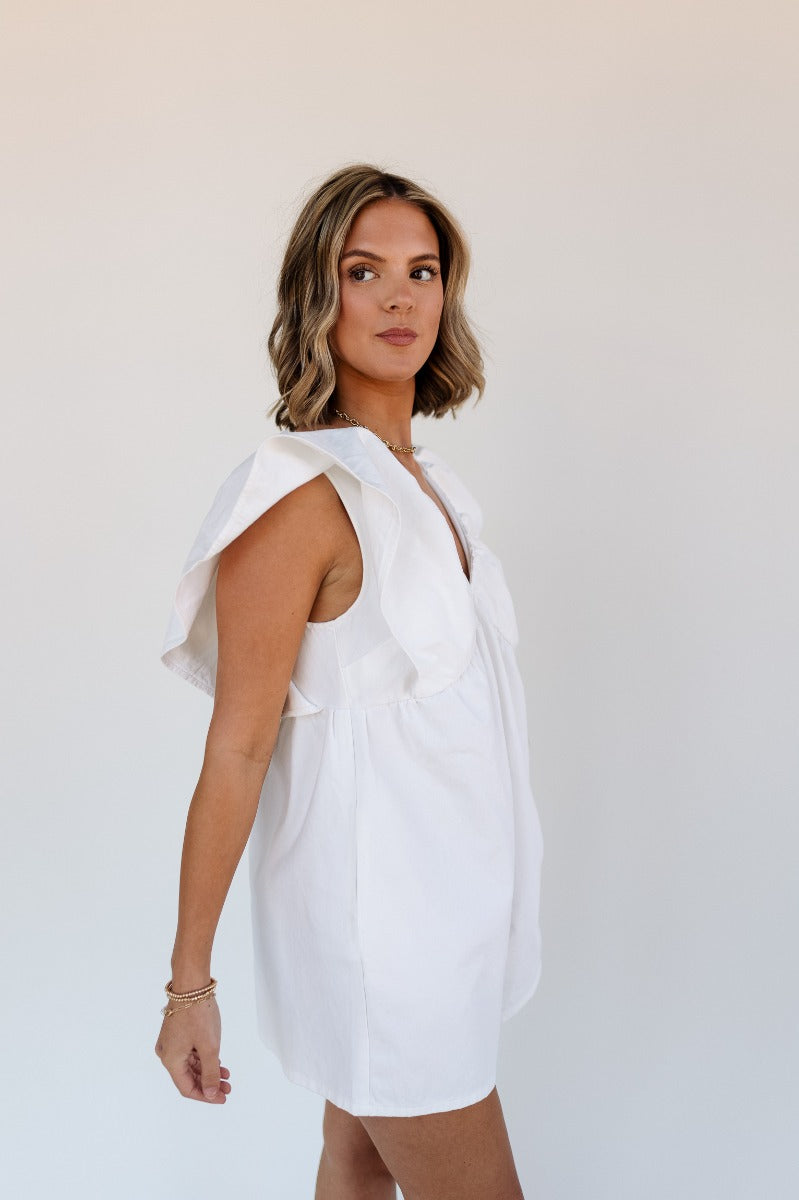Side view of model wearing the Peyton Off White Denim Sleeveless Romper that has off white cotton fabric, two front pockets, a plunge neck, ruffle straps, and a back zipper with a hook closure.