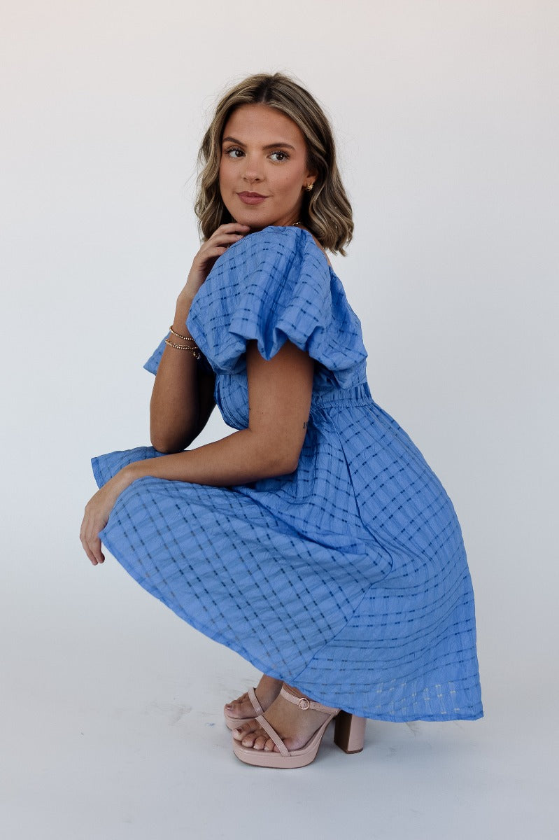 Side view of model wearing the Melanie Blue Plaid Short Sleeve Mini Dress that has blue light weight fabric, monochrome plaid print, blue lining, mini length, elastic waist, square neck and short puff sleeves.