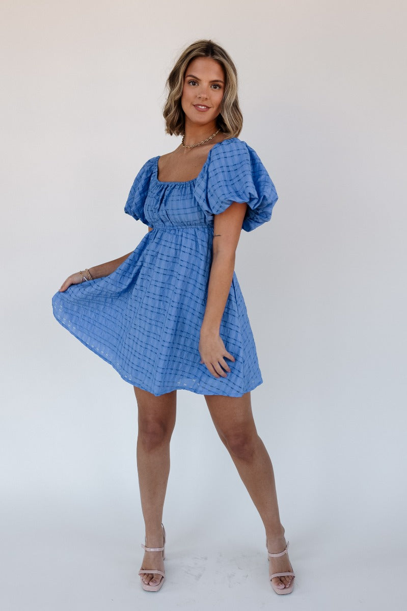 Full body front view of model wearing the Melanie Blue Plaid Short Sleeve Mini Dress that has blue light weight fabric, monochrome plaid print, blue lining, mini length, elastic waist, square neck and short puff sleeves.