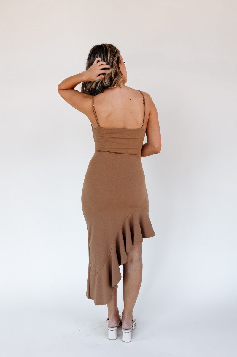 Full body back view of model wearing the Liliana Brown Asymmetrical Sleeveless Midi Dress that has light brown knit fabric, an asymetrical ruffle hem, midi length, square neckline, adjustable straps and sleeveless.
