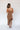 Full body back view of model wearing the Liliana Brown Asymmetrical Sleeveless Midi Dress that has light brown knit fabric, an asymetrical ruffle hem, midi length, square neckline, adjustable straps and sleeveless.