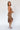 Full body side view of model wearing the Liliana Brown Asymmetrical Sleeveless Midi Dress that has light brown knit fabric, an asymetrical ruffle hem, midi length, square neckline, adjustable straps and sleeveless.