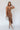 Full body front view of model wearing the Liliana Brown Asymmetrical Sleeveless Midi Dress that has light brown knit fabric, an asymetrical ruffle hem, midi length, square neckline, adjustable straps and sleeveless.