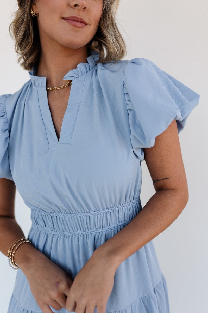 Upper front view of model wearing the Ellie Light Blue Tiered Short Sleeve Midi Dress features light blue fabric, a notched neck with ruffle trim, short puff sleeves, elastic waistband, tiered body, maxi-length, and lining. 