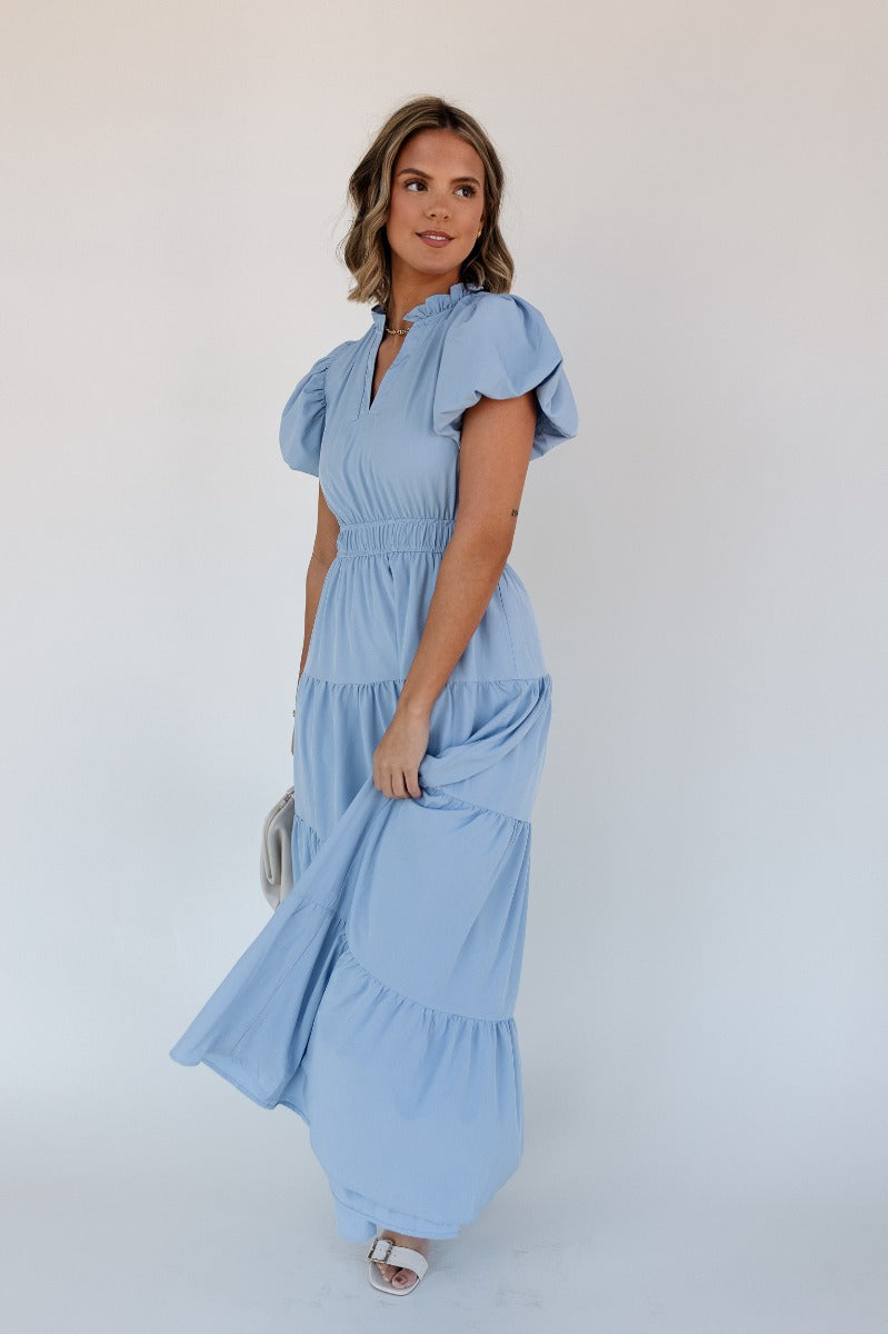 Full body front view of model wearing the Ellie Light Blue Tiered Short Sleeve Midi Dress features light blue fabric, a notched neck with ruffle trim, short puff sleeves, elastic waistband, tiered body, maxi-length, and lining. 