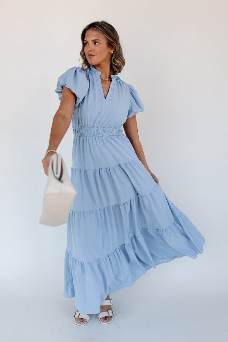 Full body front view of model wearing the Ellie Light Blue Tiered Short Sleeve Midi Dress features light blue fabric, a notched neck with ruffle trim, short puff sleeves, elastic waistband, tiered body, maxi-length, and lining. 