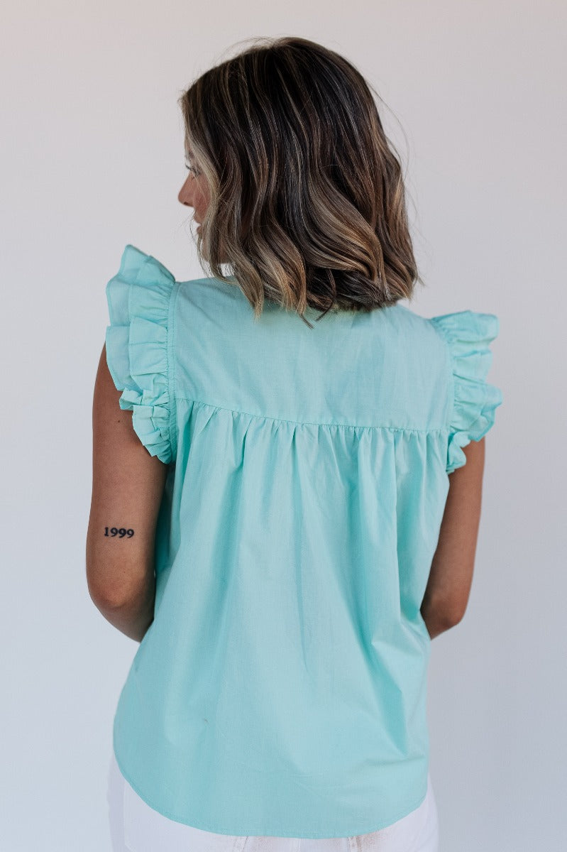 Back view of model wearing the Emory Aqua Sleeveless Ruffle Top that has aqua blue cotton fabric, a ruffled notched neckline with ties, and a sleeveless body with ruffle details.