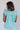 Back view of model wearing the Emory Aqua Sleeveless Ruffle Top that has aqua blue cotton fabric, a ruffled notched neckline with ties, and a sleeveless body with ruffle details.