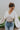 Frontal view of the Auburn Avenue Top that features an ivory colored material, a V neckline with a button closure, a long pleated sleeve, a smocked bottom with a ruffle hem, and a back tie closure.