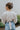 Back view of the Auburn Avenue Top that features an ivory colored material, a V neckline with a button closure, a long pleated sleeve, a smocked bottom with a ruffle hem, and a back tie closure.