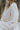 Close-up side view of model wearing the Love Always Top, which has an ivory colored material, a round neck, a 3/4 button-up front, a long sleeve, front lace detail, a babydoll design, and a flowy fit