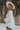 Side view of model wearing the Love Always Top, which has an ivory colored material, a round neck, a 3/4 button-up front, a long sleeve, front lace detail, a babydoll design, and a flowy fit
