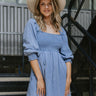 Frontal view of the Washed Away Mini Dress that features a washed blue material, a straight neckline with a ruffle trim, a 3/4 sleeve, a smocked top and back, and a mini length.