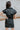 Back view of the Can't Live Without Mini Dress that features a black faux-leather material, a collar neck, a short cuffed sleeve, a button-up front, two front pockets at the chest, two front pockets at the bottom, and a mini length.
